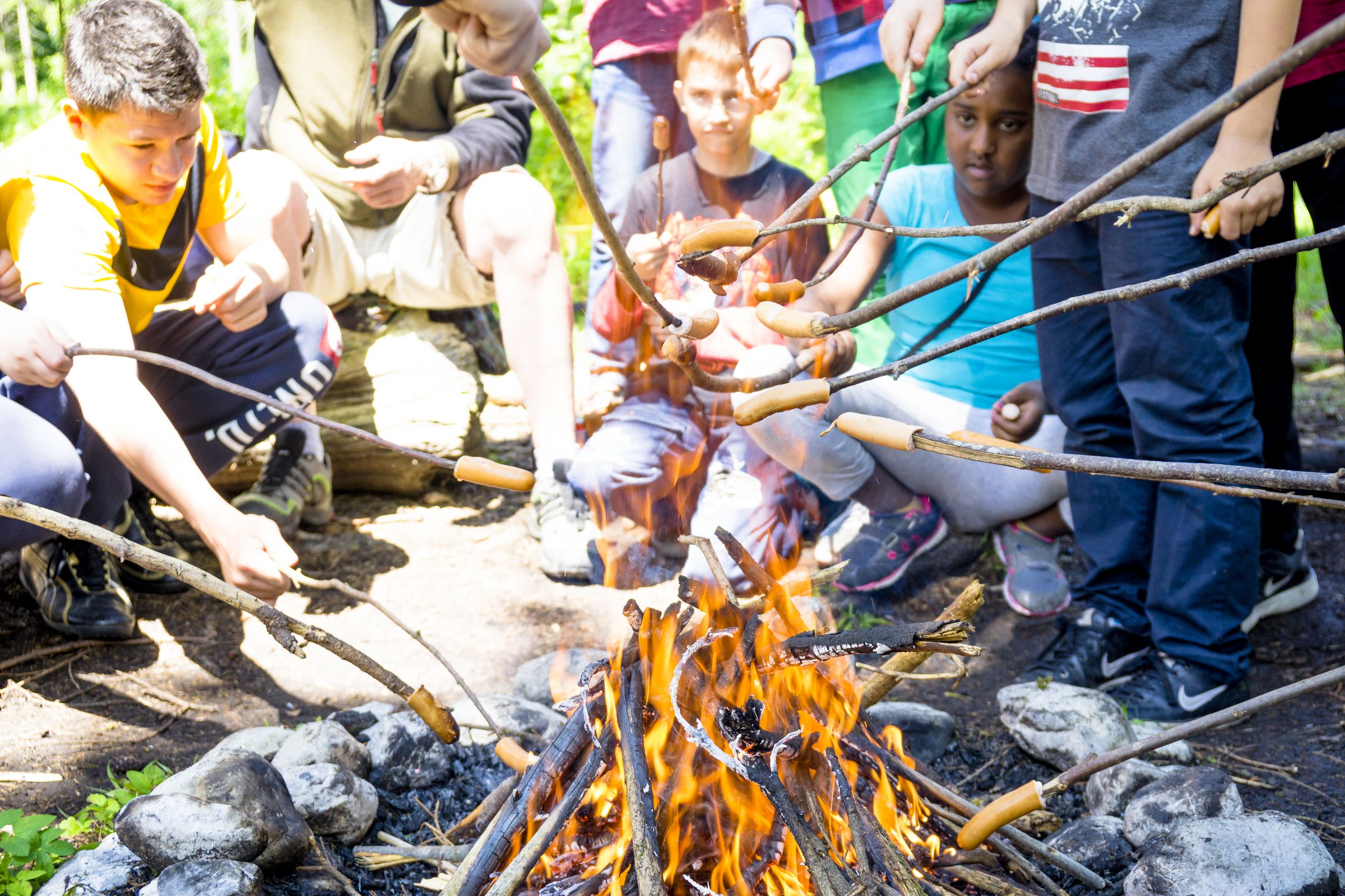Eine Gruppe Kinder sitzt um ein Lagerfeuer herum und grillt Würstchen an langen Stöcken